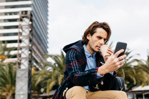 A man drinks coffee and watches the news on the phone.