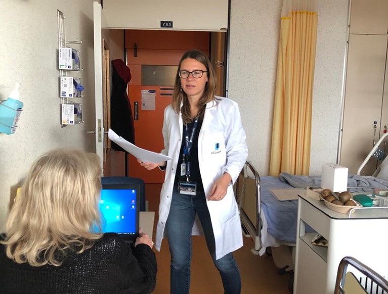 White female wearing white lab coat is standing in front of a woman seated at a computer screen.